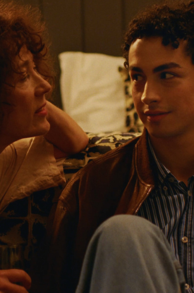 BACKSTAGE Eine Frau und ein Mann sitzen nebeneinander vor einem Bett und unterhalten sich.