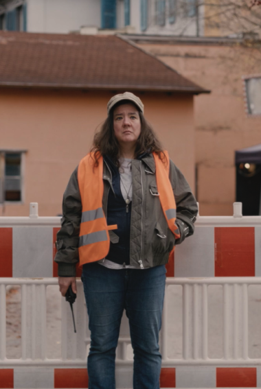 DIE ABSPERRFRAU - TRAUMJOB FILMBRANCHE Eine Frau in orangener Warnweste steht vor einer Straßenabsperrung.