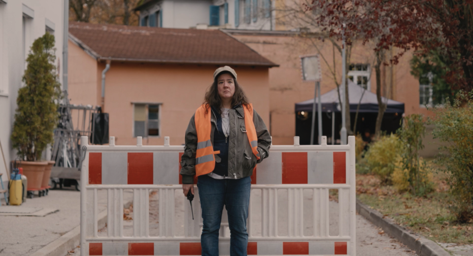 Eine Frau in orangener Warnweste steht vor einer Straßenabsperrung. Eine Frau in orangener Warnweste steht vor einer Straßenabsperrung.