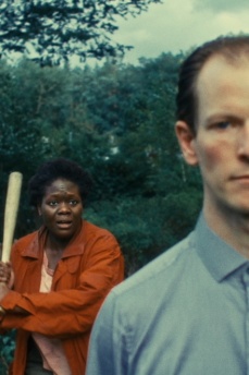 Im Vordergrund steht ein Mann, im Hintergrund bewegt sich eine Frau mit Baseballschläger in der Hand auf ihn zu.