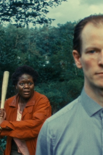 DER TOTALE TRAUM Im Vordergrund steht ein Mann, im Hintergrund bewegt sich eine Frau mit Baseballschläger in der Hand auf ihn zu.