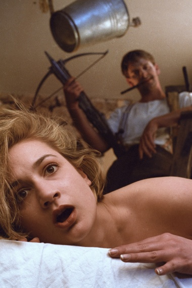 Im Vordergrund liegt eine Frau im Bett, im Hintergrund ist ein Mann zu sehen, der eine Armbrust in der Hand hält.