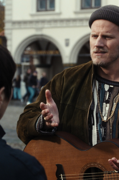 THE FOLKS Ein Mann mit Mütze hält eine Gitarre in seiner Hand und unterhält sich in einer Innenstadt mit einer Frau, die ihm gegenüber steht.