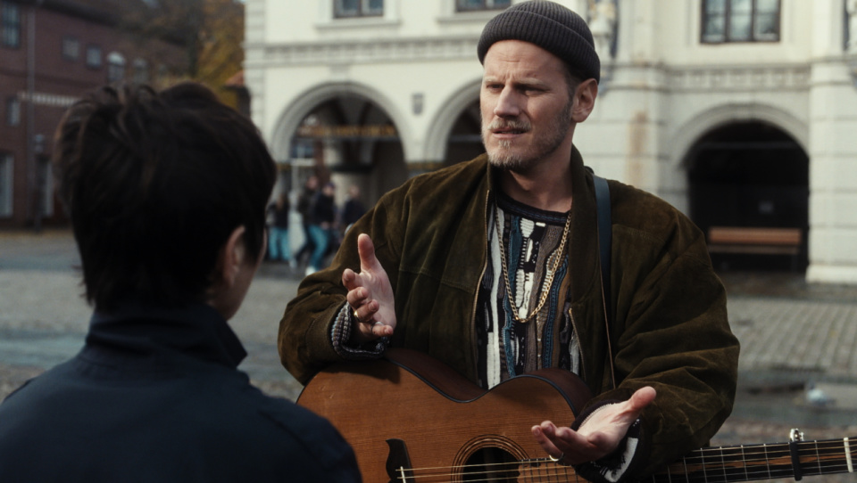 Ein Mann mit Mütze hält eine Gitarre in seiner Hand und unterhält sich in einer Innenstadt mit einer Frau, die ihm gegenüber steht. Ein Mann mit Mütze hält eine Gitarre in seiner Hand und unterhält sich in einer Innenstadt mit einer Frau, die ihm gegenüber steht.
