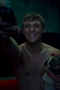 Ein Mann sitzt grinsend oberkörperfrei in einem Raum. Er hält eine Fernbedienung in der Hand und hat auf der anderen Seite einen Mops auf Arm.