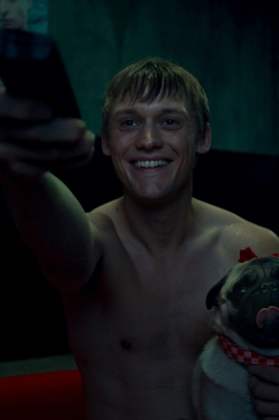 Ein Mann sitzt grinsend oberkörperfrei in einem Raum. Er hält eine Fernbedienung in der Hand und hat auf der anderen Seite einen Mops auf Arm.