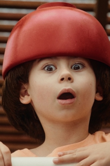 COUPE AU BOL Ein Kind schaut erschrocken über ein Geländer. Es trägt eine rote Kopfbedeckung.