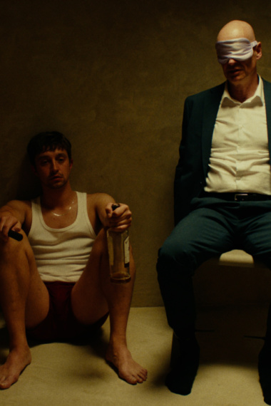 Zwei Männer sitzen in einer Dusche. Einer von ihnen trägt einen Anzug und eine Augenbinde, der andere sitzt links von ihm in Unterwäsche und hält eine Schnapsflasche in der Hand.