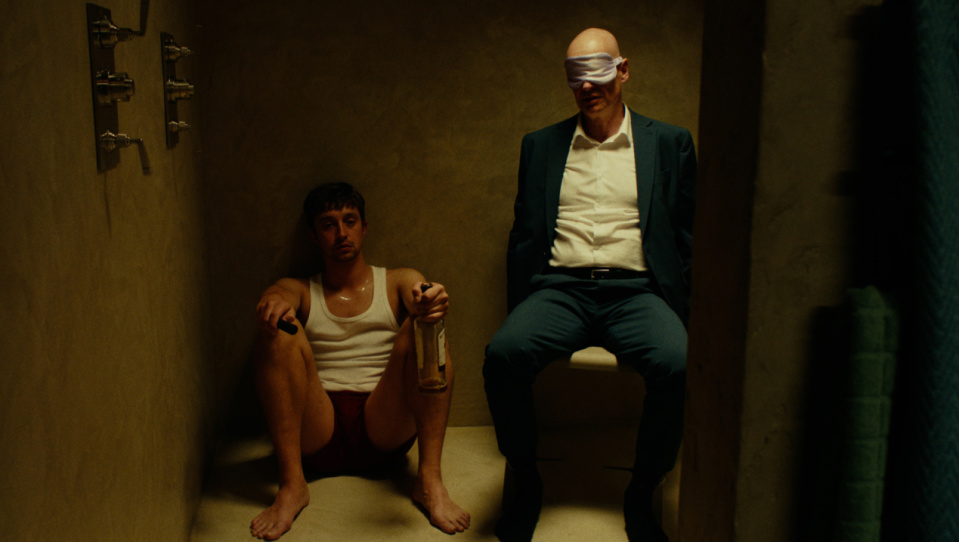 Zwei Männer sitzen in einer Dusche. Einer von ihnen trägt einen Anzug und eine Augenbinde, der andere sitzt links von ihm in Unterwäsche und hält eine Schnapsflasche in der Hand.