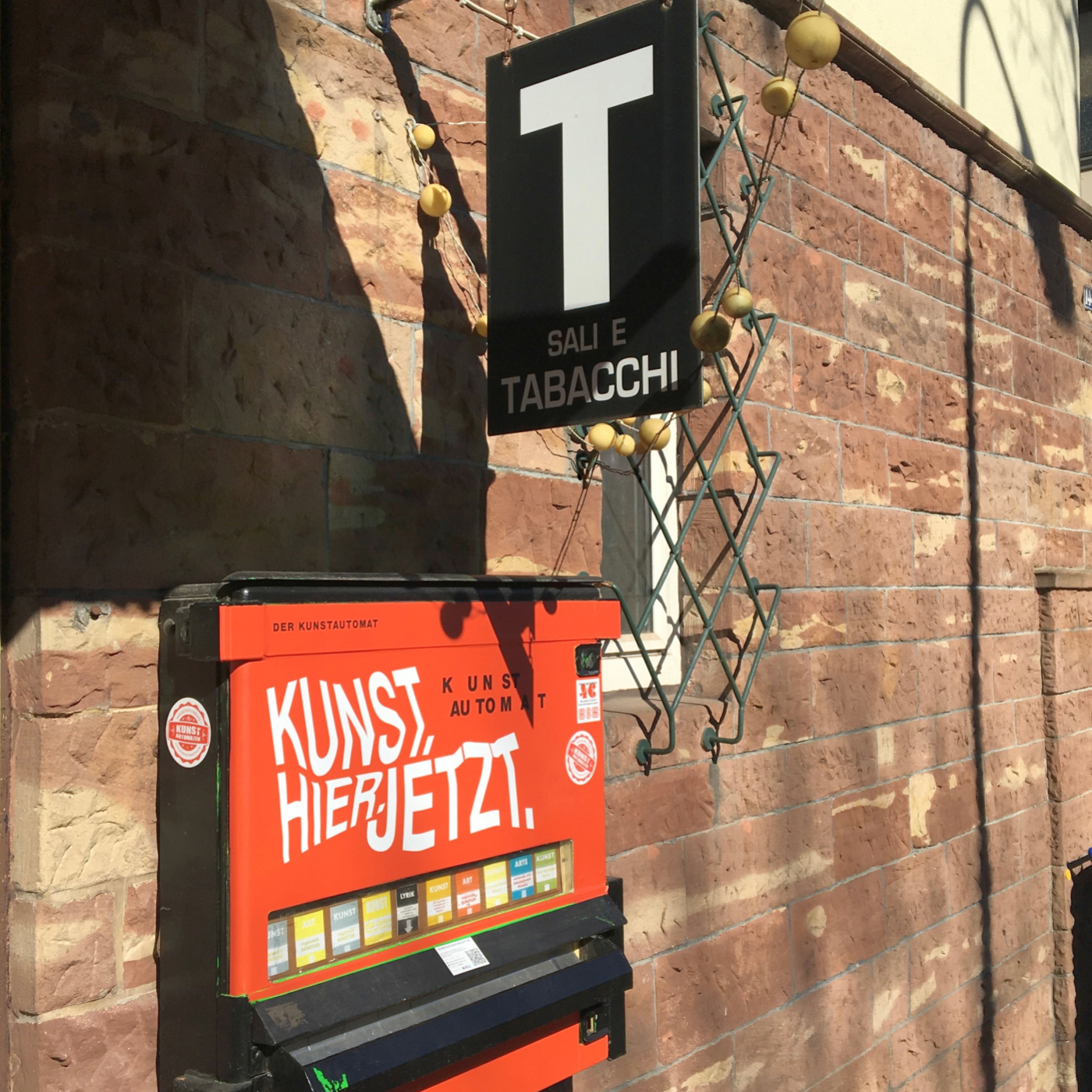 Ein roter Automat mit der Aufschrift Kunst. Hier. Jetzt. und ein Schild mit der Aufschrift Slai e Tabacchi an einer Hauswand