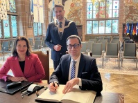 Ein Mann und eine Frau sitzen an einem Tisch in einem Festsaal. Der Mann schreibt etwas in ein Buch. Hinter den beiden steht ein weiterer Mann. Alle drei blicken in die Kamera.