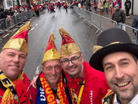 Ein Selfie zeigt vier Männer in Karnevalskleidung und Narrenkappen vor der abgesperrten Umzugsstrecke.