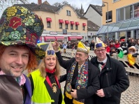 Vier Personen mit Narrenkappen posieren für ein Selfie vor einer Menschenmenge am Straßenrand.