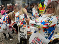 Mehrere Frauen in bunten Jacken und mit Tragetaschen posieren vor einer großen Zuschauermenge am Straßenrand.