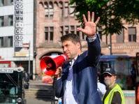 Mann im Anzug spricht mit rotem Megafon und hebt die Hand. Im Hintergrund stehen mehrere Personen und Fahrzeuge.