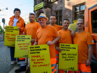 Drei Personen stehen mit Schildern vor einem orangefarbenen Kommunalfahrzeug, im Hintergrund ist ein historisches Gebäude zu sehen.