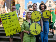 Vier Personen halten gelbe Uhrensymbole, daneben ist ein Protestplakat zu sehen. Im Hintergrund stehen weitere Teilnehmende.