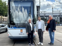 Drei Personen stehen vor einer Straßenbahn auf Gleisen, im Hintergrund sind Oberleitungen und ein Betriebshof zu sehen.