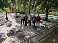 Fünf Personen stehen auf einer Sandfläche mit Liegestühlen und Tischen neben einem Freibad, umgeben von Bäumen und Sonnenschirmen.