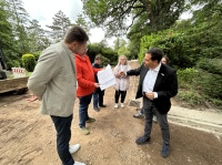 Personengruppe bespricht auf einem unbefestigten Weg Pläne für Bauarbeiten in einer grünen Friedhofsanlage.