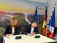 Zwei Personen unterzeichnen Dokumente an einem Tisch vor Flaggen und einer Landschaftsgrafik an der Wand.