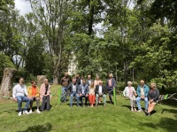 Personengruppe sitzt und steht auf einer Sitz- und Kletteranlage auf einer Wiese, umgeben von hohen Bäumen.