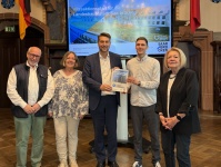 Fünf Personen präsentieren in einem historischen Saal den Hitzeaktionsplan vor einem Bildschirm und den Flaggen Deutschlands und Frankreichs.