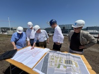 Fünf Personen mit Schutzhelmen betrachten auf einer Baustelle Pläne, die auf einem Tisch ausgebreitet sind.