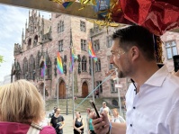 Blick von einem festlich geschmückten Wagen auf ein historisches Gebäude mit Regenbogenflaggen. Menschen stehen auf dem Platz.