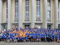 Gruppe von Menschen in blauen und orangefarbenen Laufshirts vor einem Gebäude mit Säulen. Viele winken in die Kamera.
