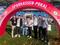 Sieben Menschen posieren mit einem Pokal unter einem roten Torbogen mit der Aufschrift „Sparkassen-Pokal“ in einem Stadion.