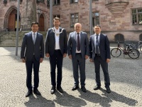 Vier Männer in Anzügen stehen vor einem historischen Gebäude zwischen Fahnenmasten auf einem gepflasterten Platz.