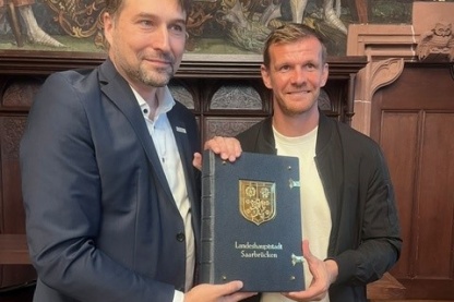 Zwei Männer halten gemeinsam ein Gästebuch mit dem Wappen und der Aufschrift „Landeshauptstadt Saarbrücken“.