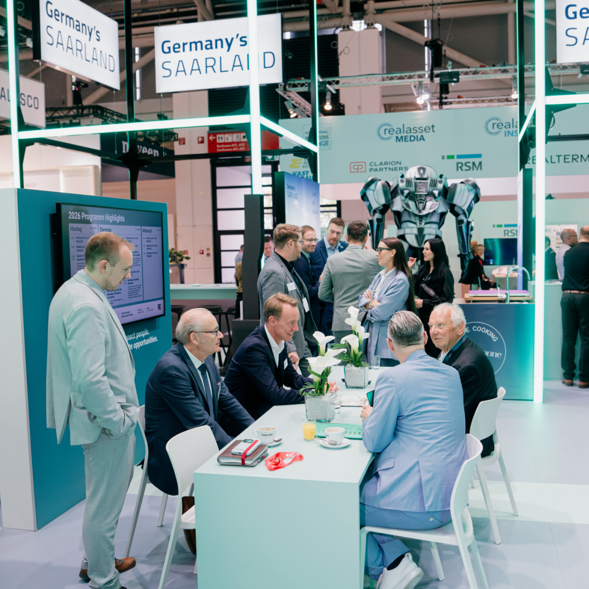 Am Messestand Germany's Saarland sitzen vier Männer an einem Besprechungstisch, ein Mann steht daneben. Im Hintergrund sind weitere Personen, die sich unterhalten.