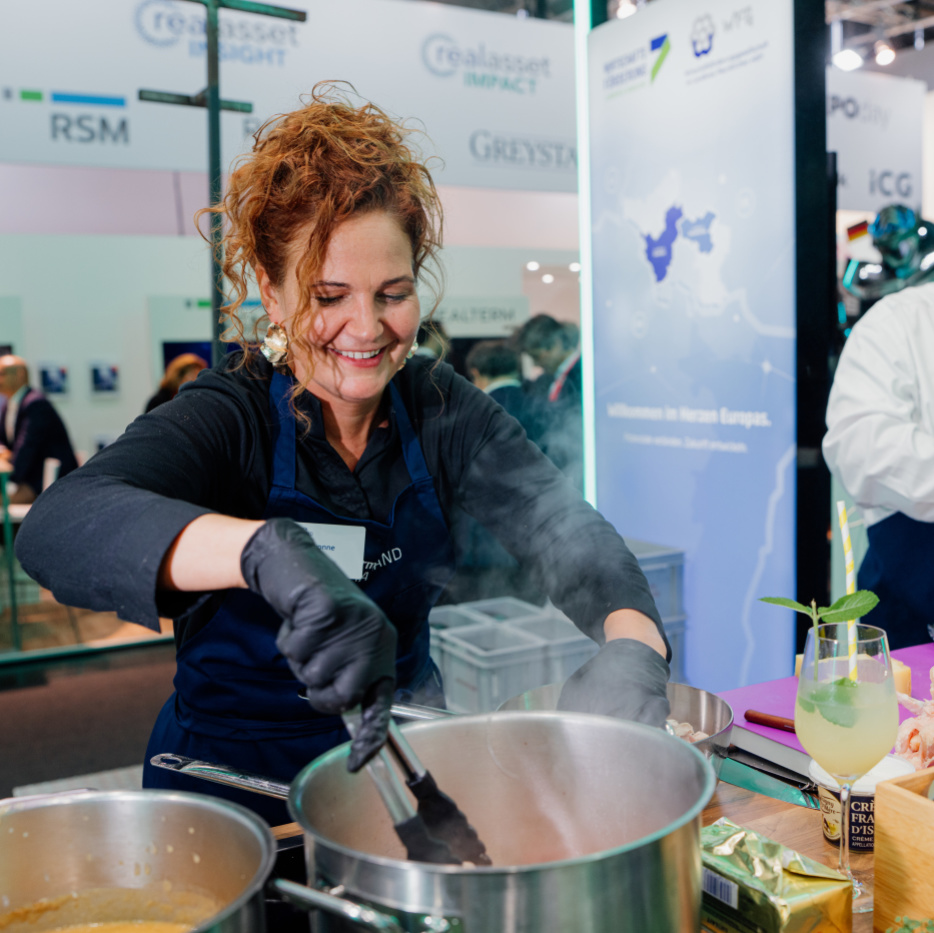 Eine lachende Frau greift mit einer Zange in einen dampfenden Kochtopf. Rechts neben ihr schneidet ein Mann eine Zwiebel mit einem großen Messer.